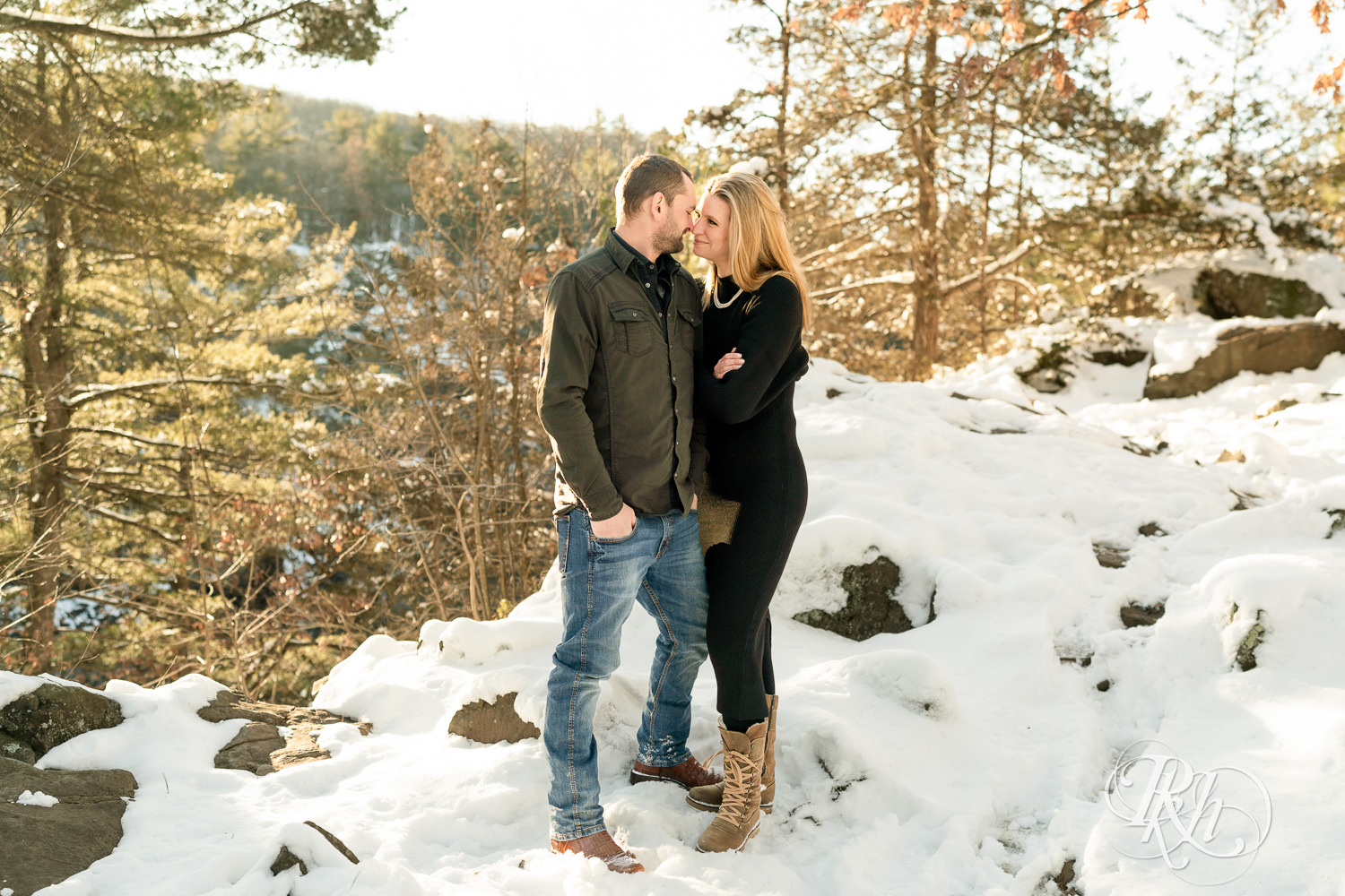 Man in jeans and woman in black dress smile in the snow at sunrise during Taylor's Falls engagement photography session.