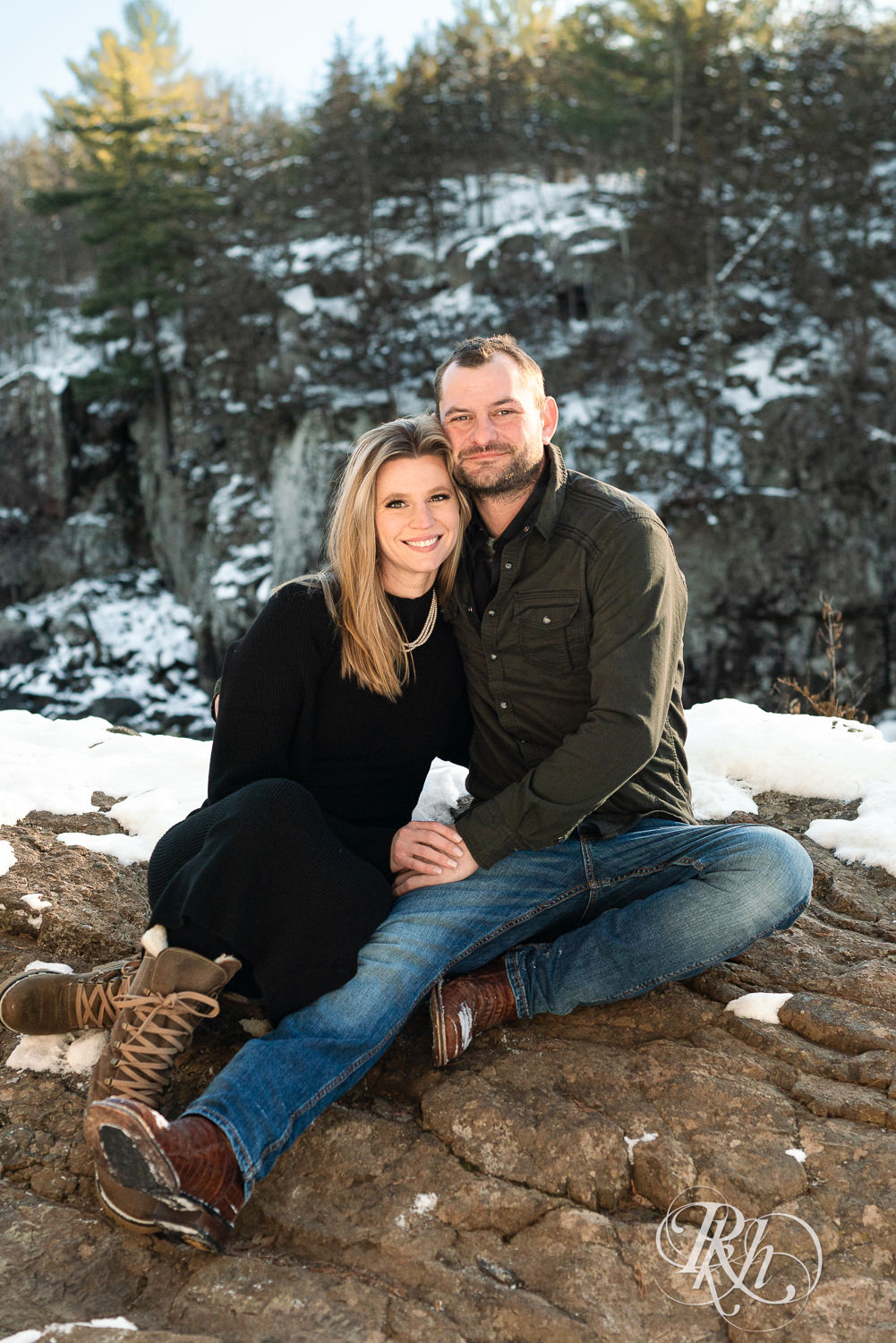 Man in jeans and woman in black dress smile on a rock at sunrise during Taylor's Falls engagement photography session.