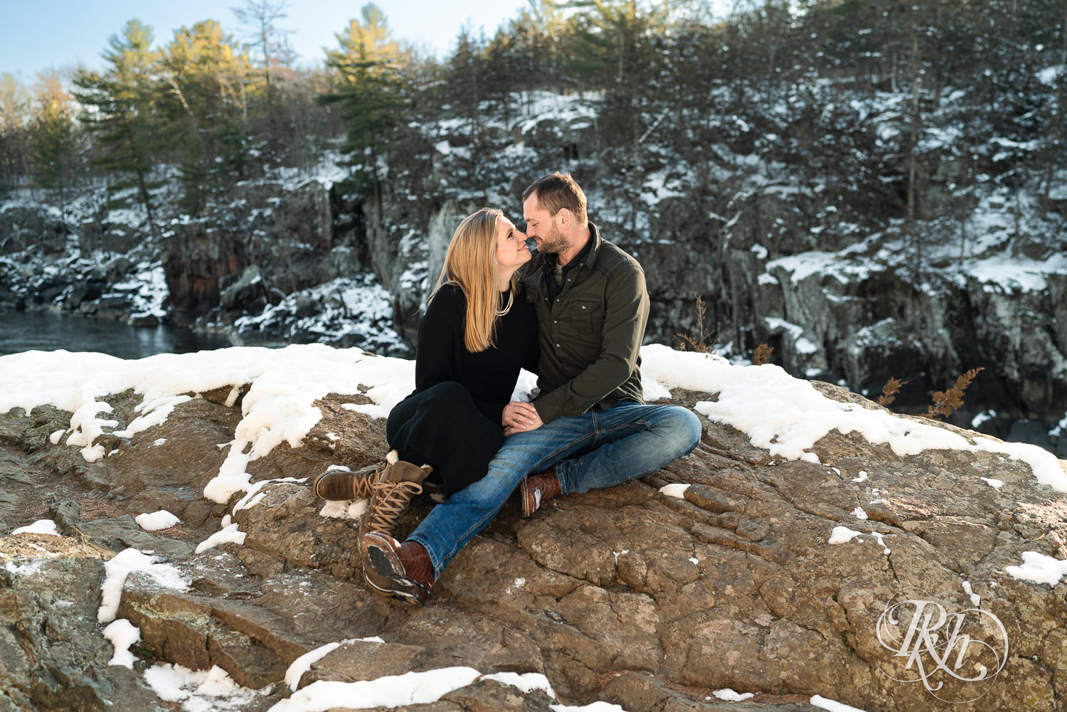 Man in jeans and woman in black dress smile on a rock at sunrise during Taylor's Falls engagement photography session.