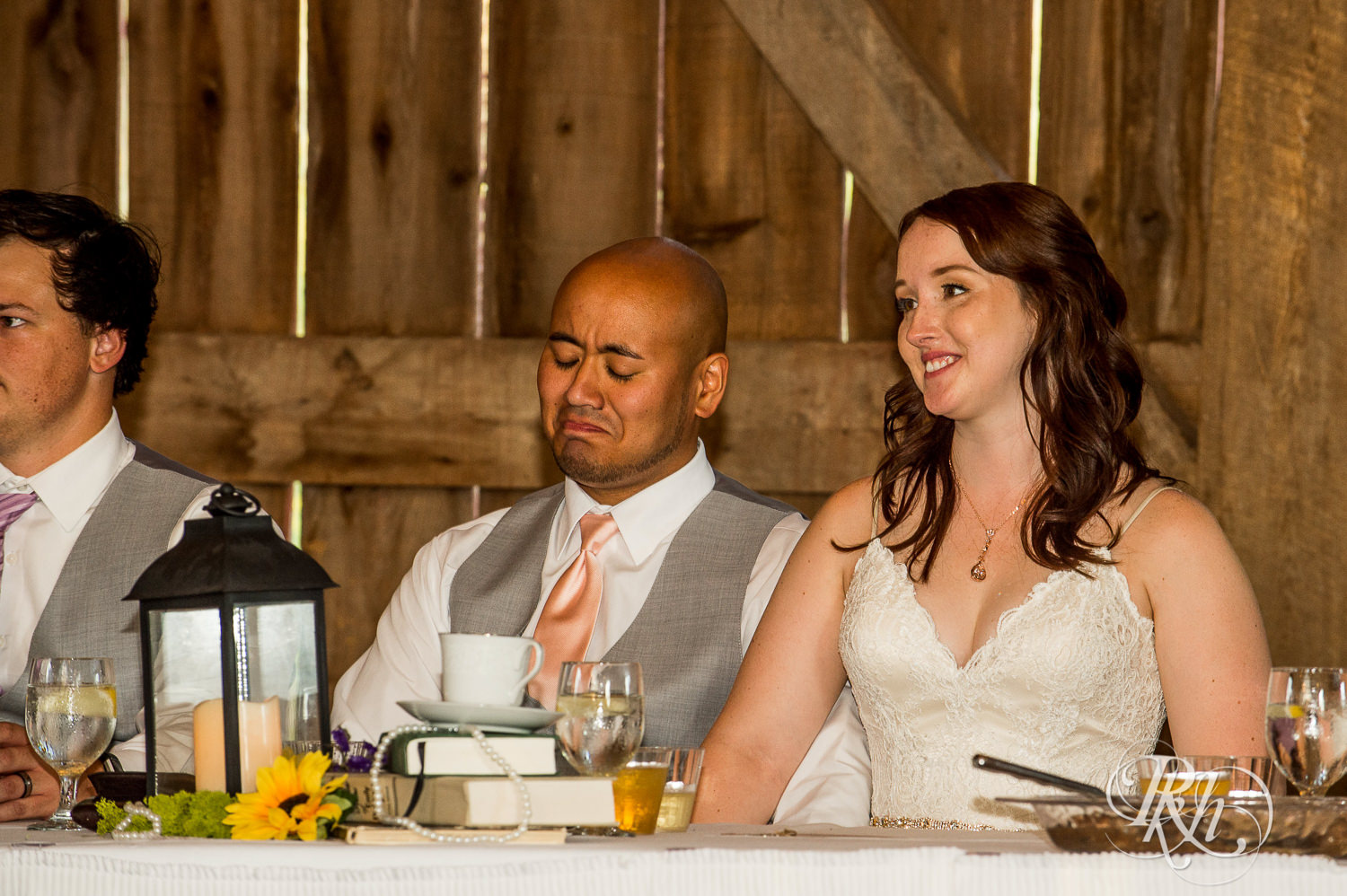 Bride and Asian groom smile during wedding reception speeches at Birch Hill Barn in Glenwood City, Wisconsin.