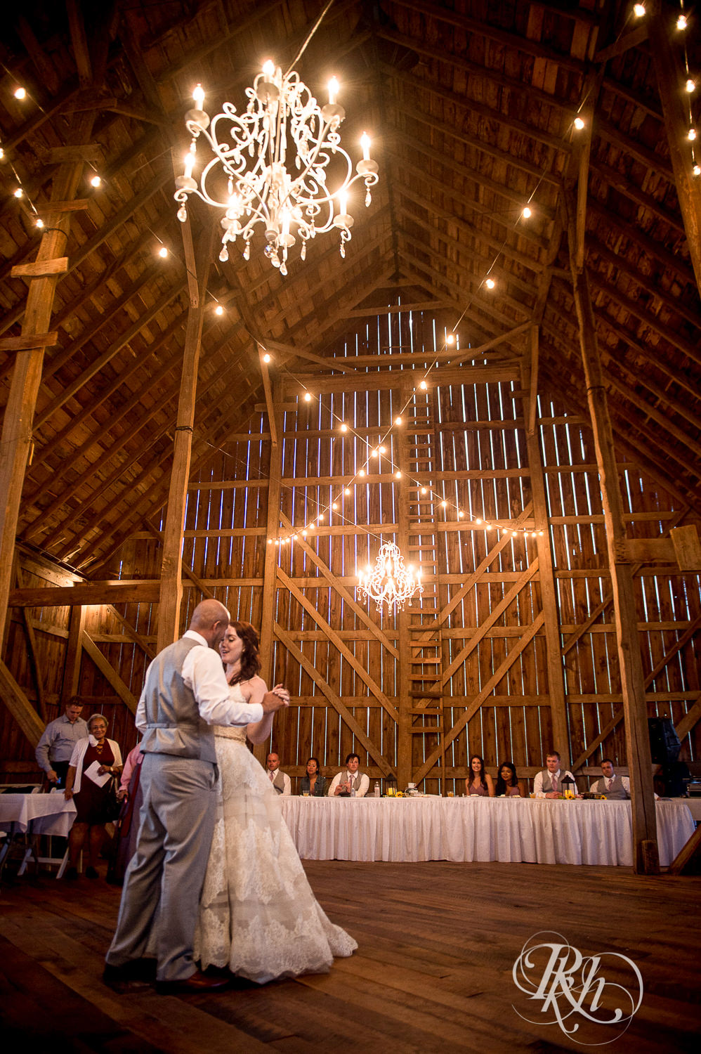 Bride and Asian groom dance under a chandelier during wedding reception at Birch Hill Barn in Glenwood City, Wisconsin.