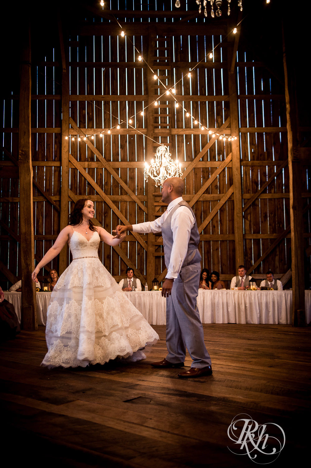Bride and Asian groom dance under a chandelier during wedding reception at Birch Hill Barn in Glenwood City, Wisconsin.
