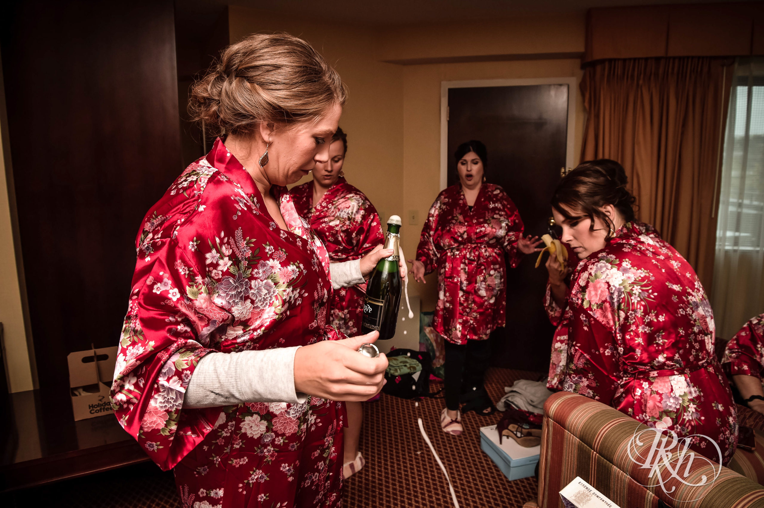Bridesmaids pop open a bottle of champagne in hotel room. 