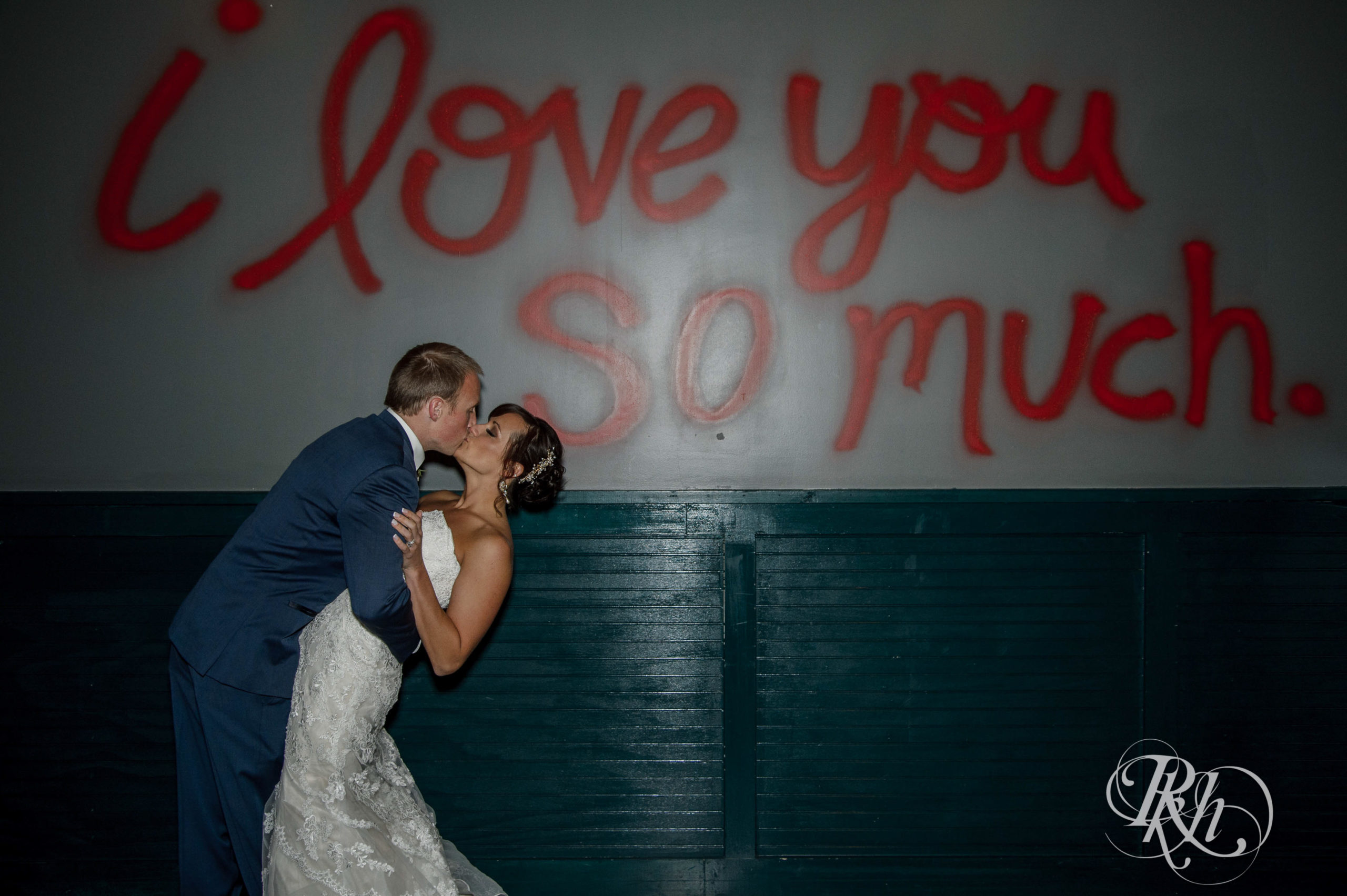 Bride and groom kiss under I LOVE YOU SO MUCH sign at Punch Bowl Social on wedding day in Minneapolis, Minnesota.