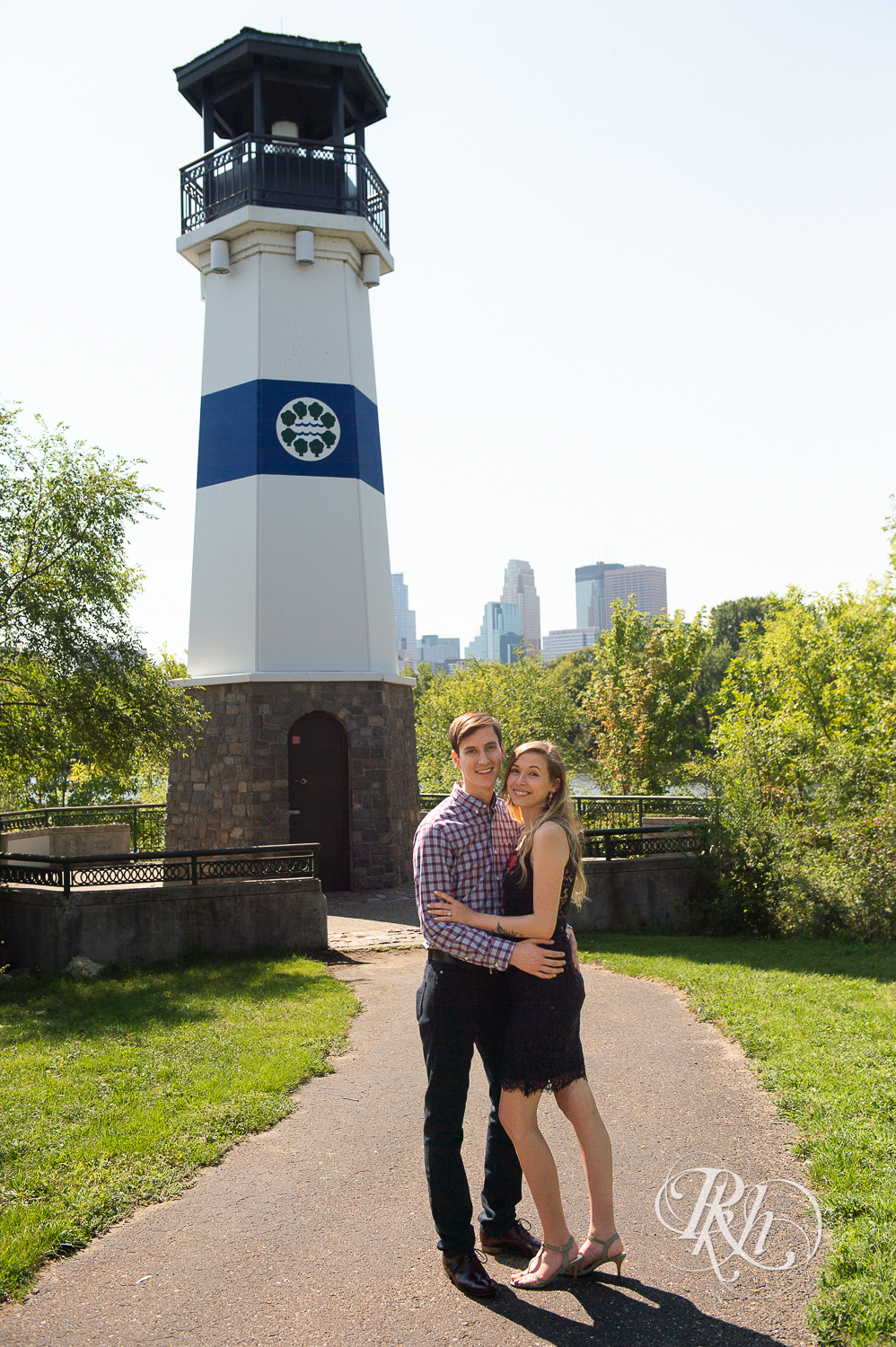 Man and woman in black lace dress smile in front of lighthouse during summer Boom Island Park engagement photos in Minneapolis, Minnesota.