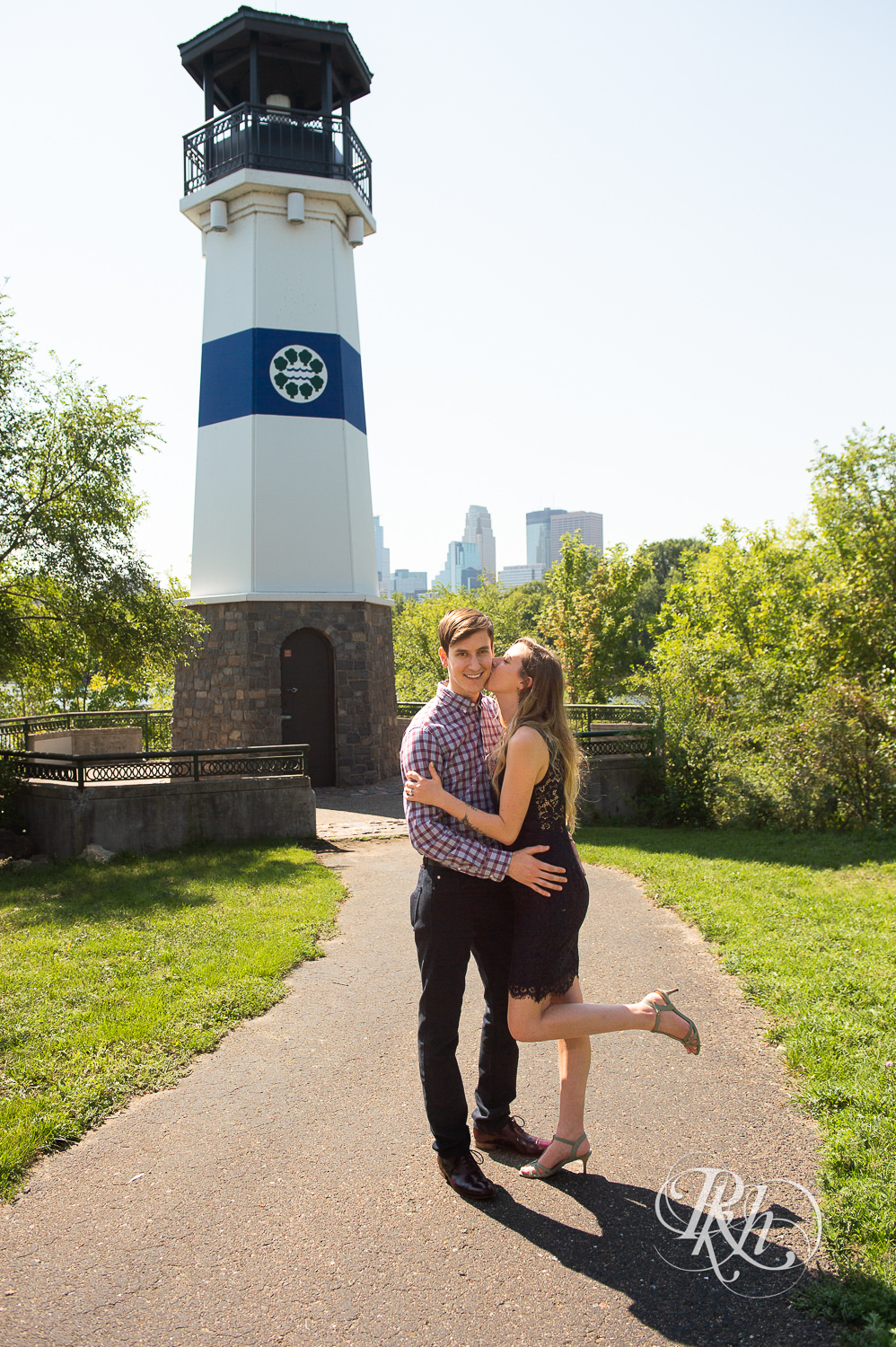 Man and woman in black lace dress smile in front of lighthouse during summer Boom Island Park engagement photos in Minneapolis, Minnesota.