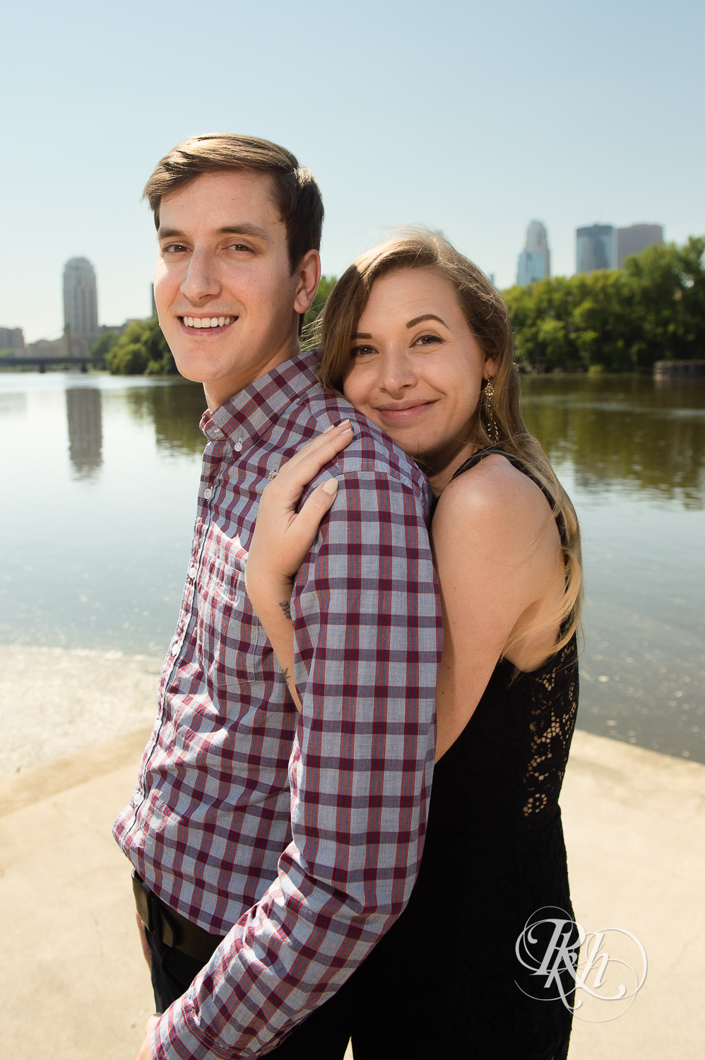 Man and woman in black lace dress smile in front of river during summer Boom Island Park engagement photos in Minneapolis, Minnesota.
