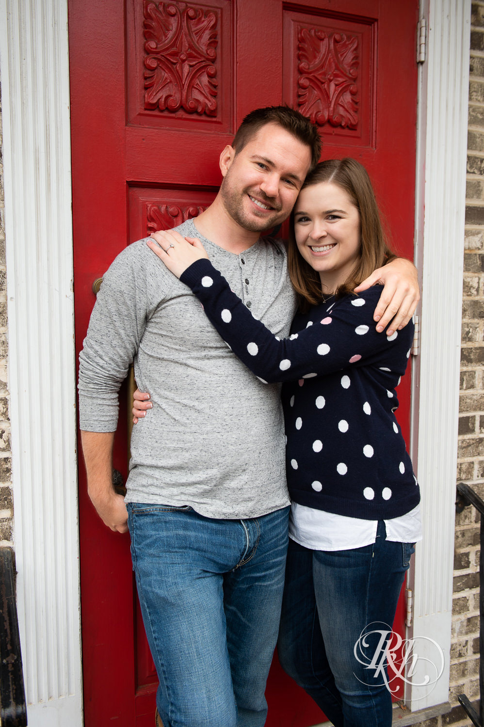 Man and woman smile against red door on rainy day in Excelsior, Minnesota.