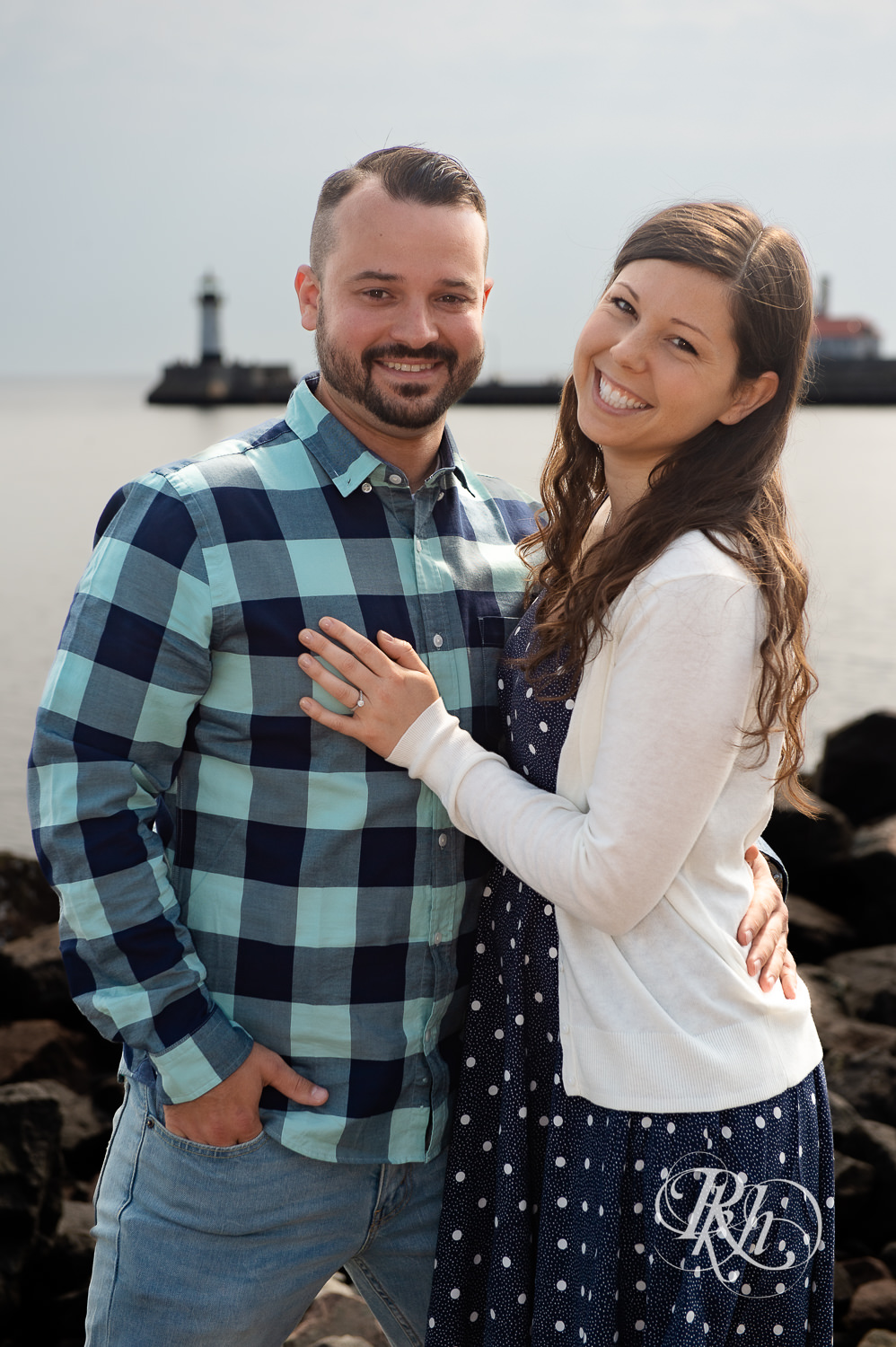 Man and woman smile with lighthouse in background during Canal Park engagement photos in Duluth, Minnesota.
