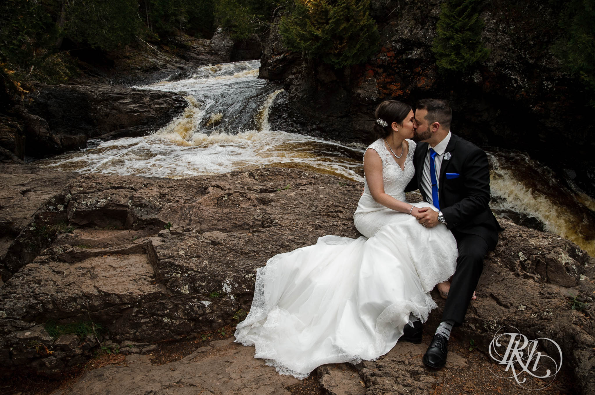 Bride and groom kiss on cliff at the bottom of a waterfall in North Shore in Minnesota.