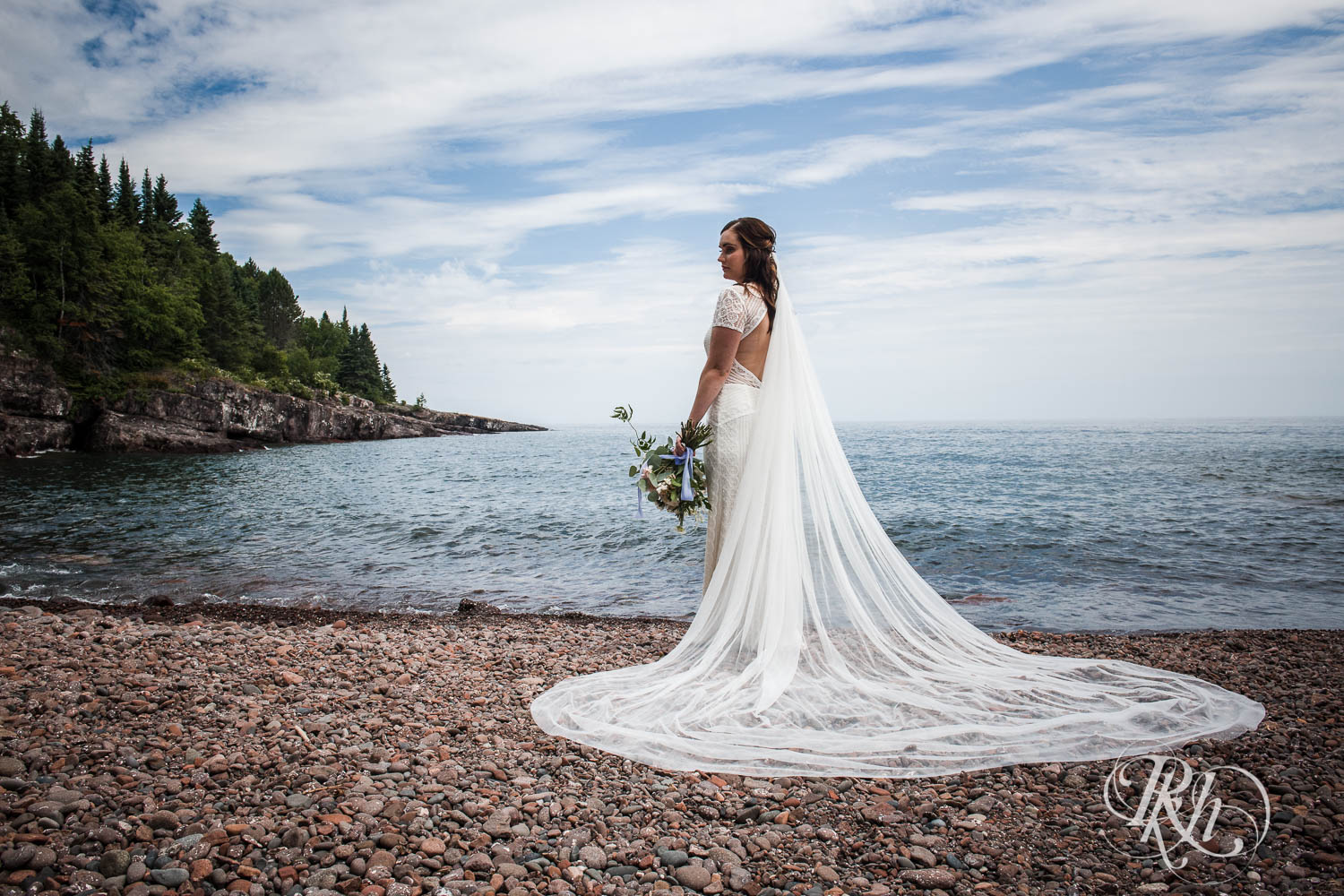 Bride with long veil and flowers stands on beach in Bluefin Bay in Tofte, Minnesota.