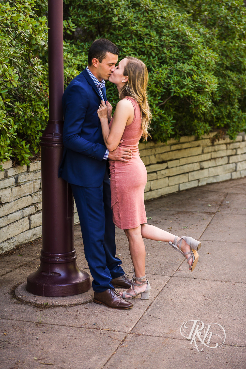 Man in blue suit and woman in pink dress kiss against pole in Centennial Lakes Park in Edina, Minnesota.