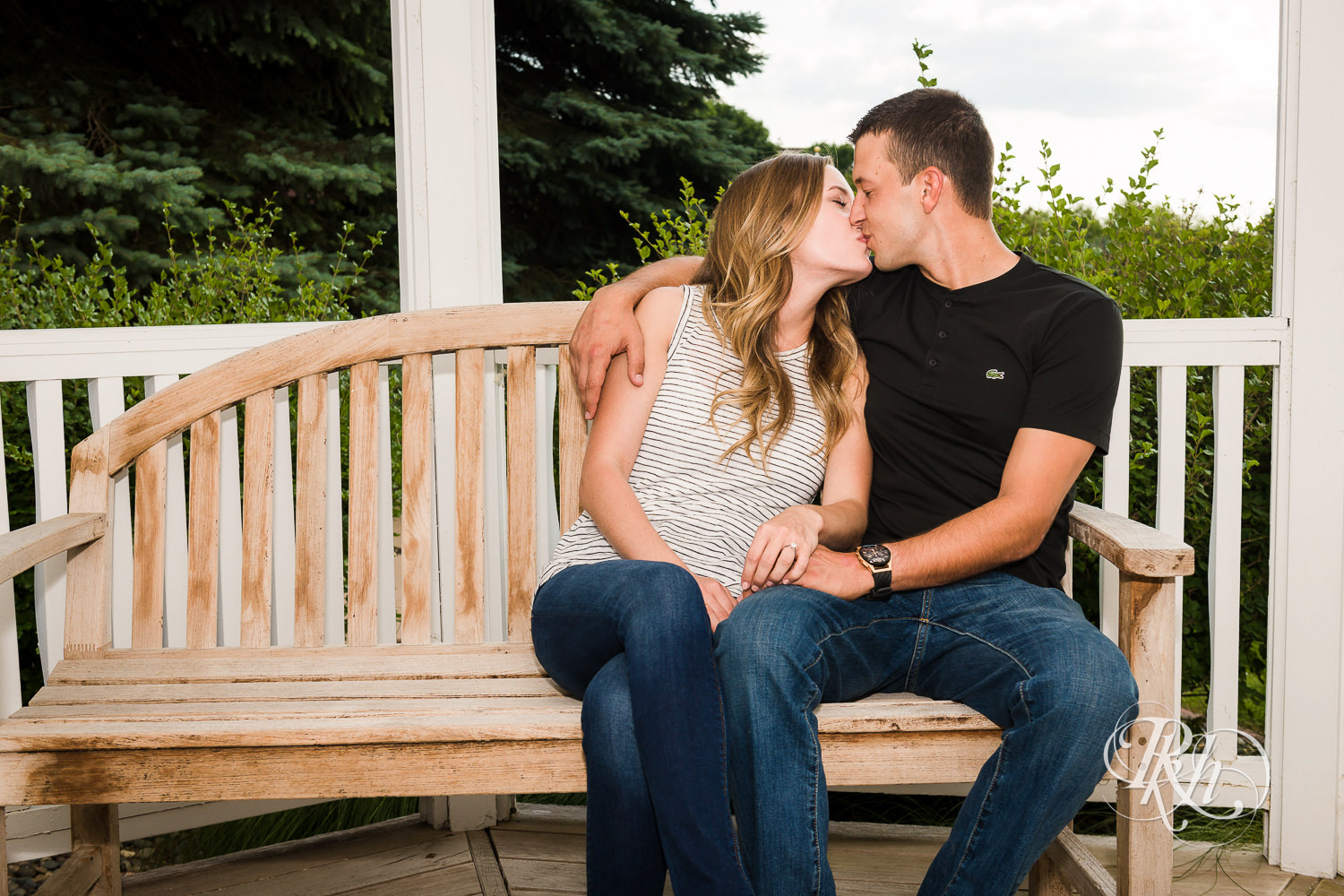 Man and woman in T-shirts and jeans kiss on bench in Centennial Lakes Park in Edina, Minnesota.