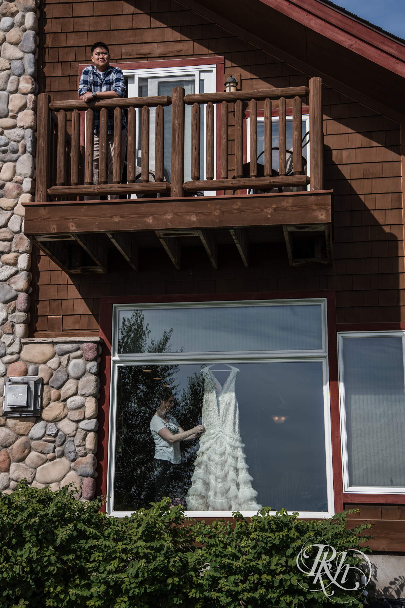 Groom standing on balcony while bride fixes wedding dress below at Grand Superior Lodge in Two Harbors, Minnesota.