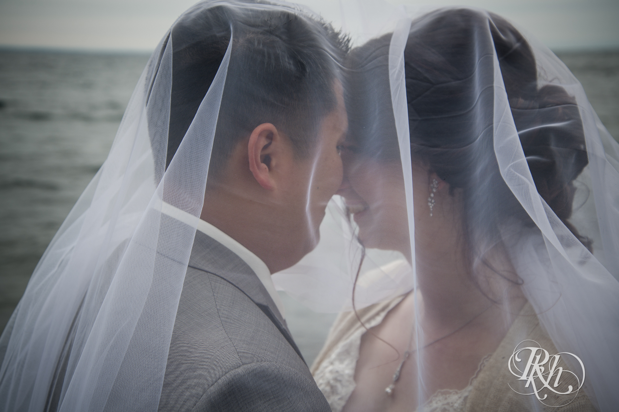 Bride and groom smile under veil in front of Lake Superior at Grand Superior Lodge in Two Harbors, Minnesota.