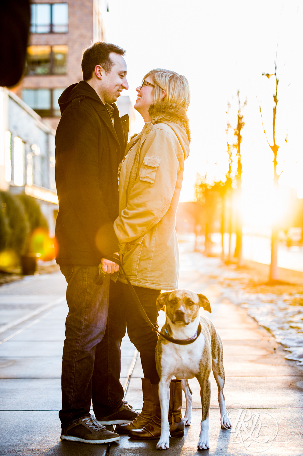 Man and woman smile while walking their dog during sunrise engagement photography in Minneapolis, Minnesota.