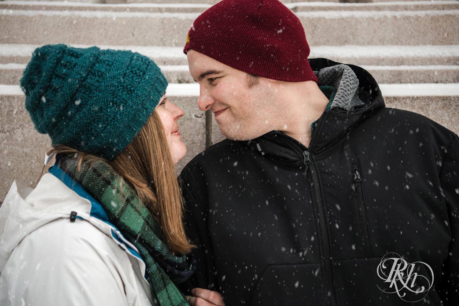 Man and woman smile in the falling snow during engagement photography in Saint Paul, Minnesota.