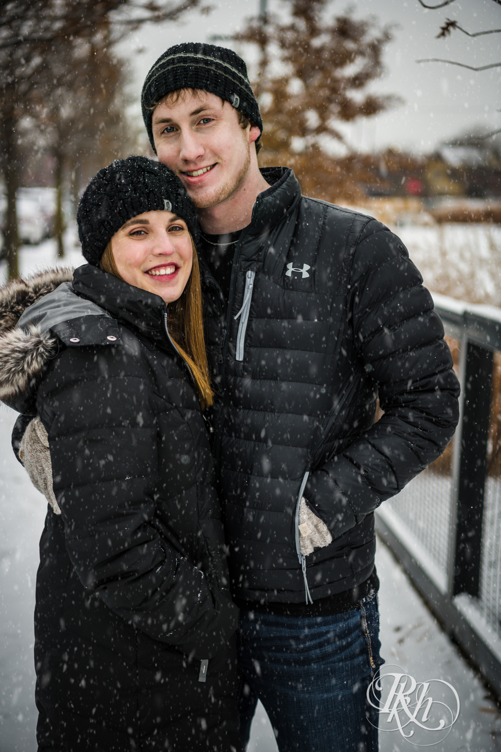 Man and woman smile in the falling snow during winter engagement photos in Saint Paul, Minnesota.