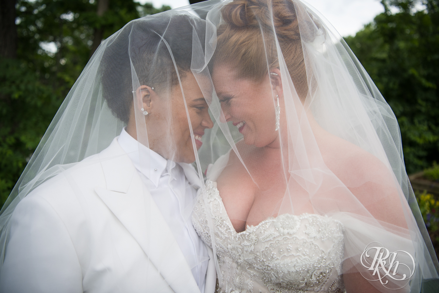 Black lesbian bride and white bride smile under veil at Leopold's Mississippi Gardens in Brooklyn Park, Minnesota.