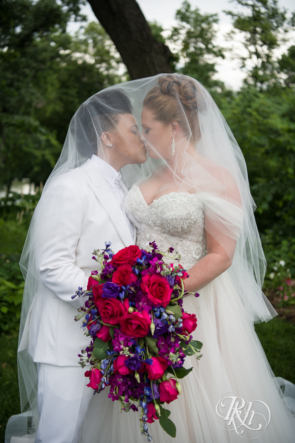 Black lesbian bride and white bride kiss under veil at Leopold's Mississippi Gardens in Brooklyn Park, Minnesota.