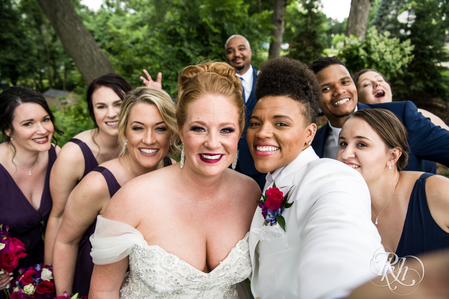 Black lesbian bride and white bride smile with wedding party at Leopold's Mississippi Gardens in Brooklyn Park, Minnesota.