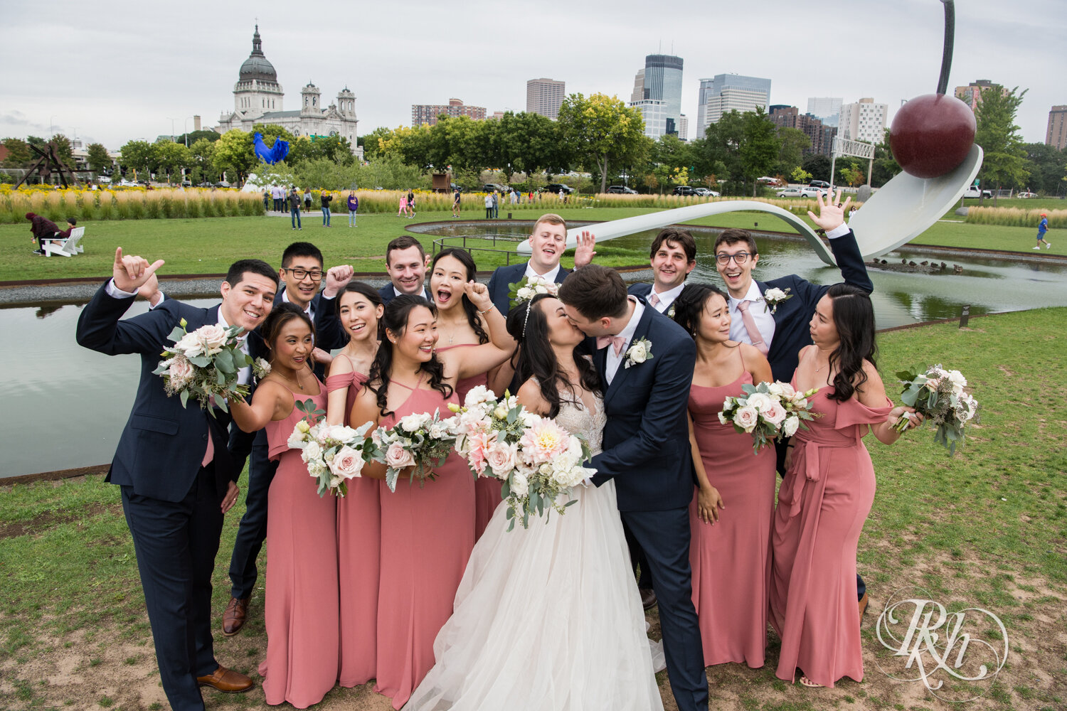 Asian bride and groom kiss in front of wedding party in Sculpture Garden in Minneapolis, Minnesota.