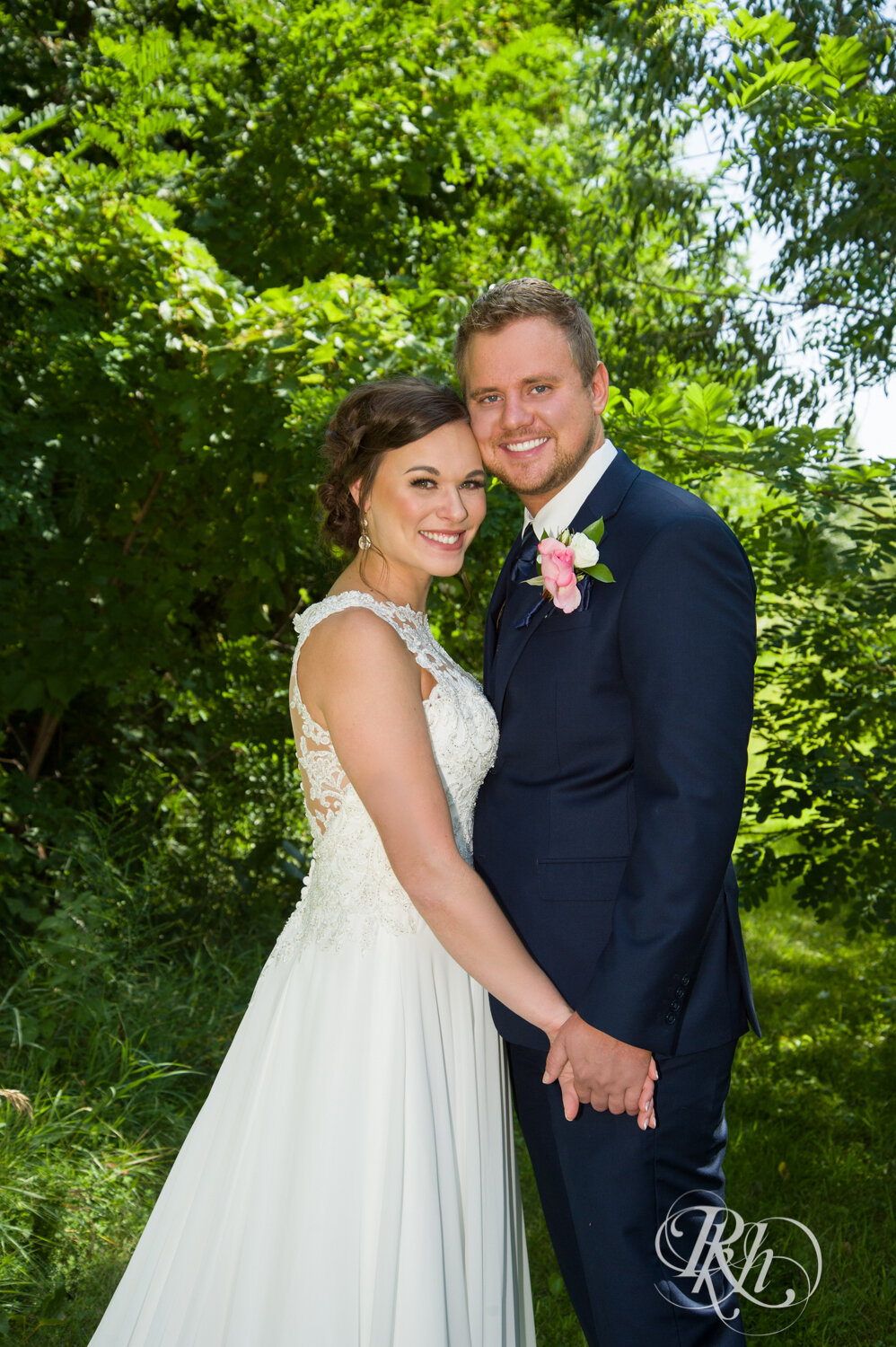 Bride and groom smile between trees on sunny wedding day at Crown Room in Rogers, Minnesota.