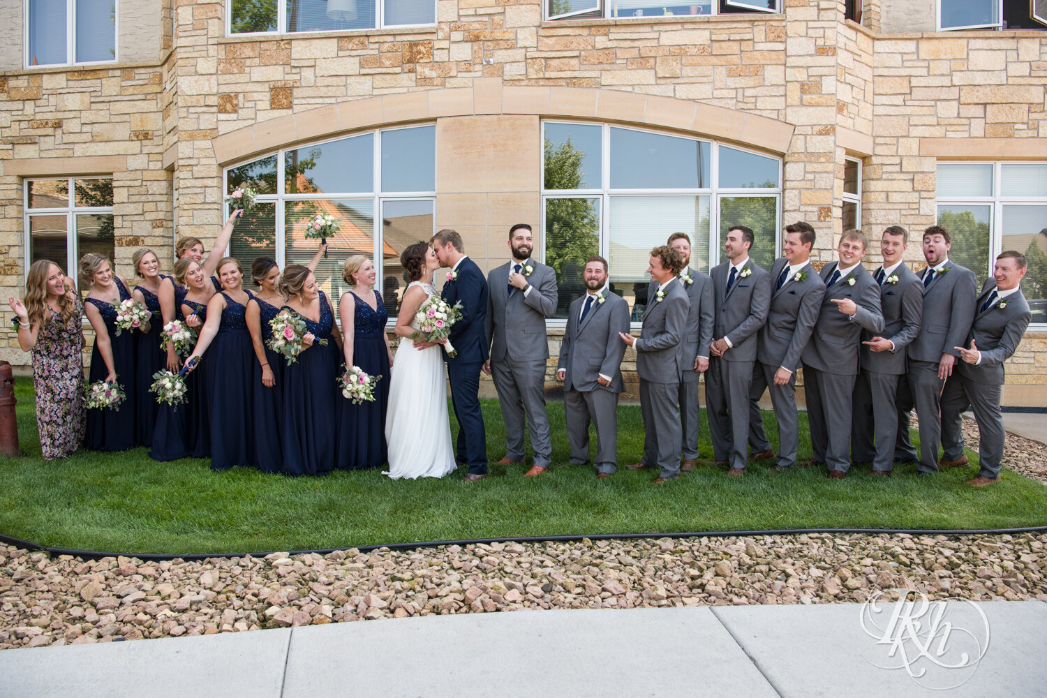 Large wedding party in blue dresses and grey suits smile at Crown Room in Rogers, Minnesota.