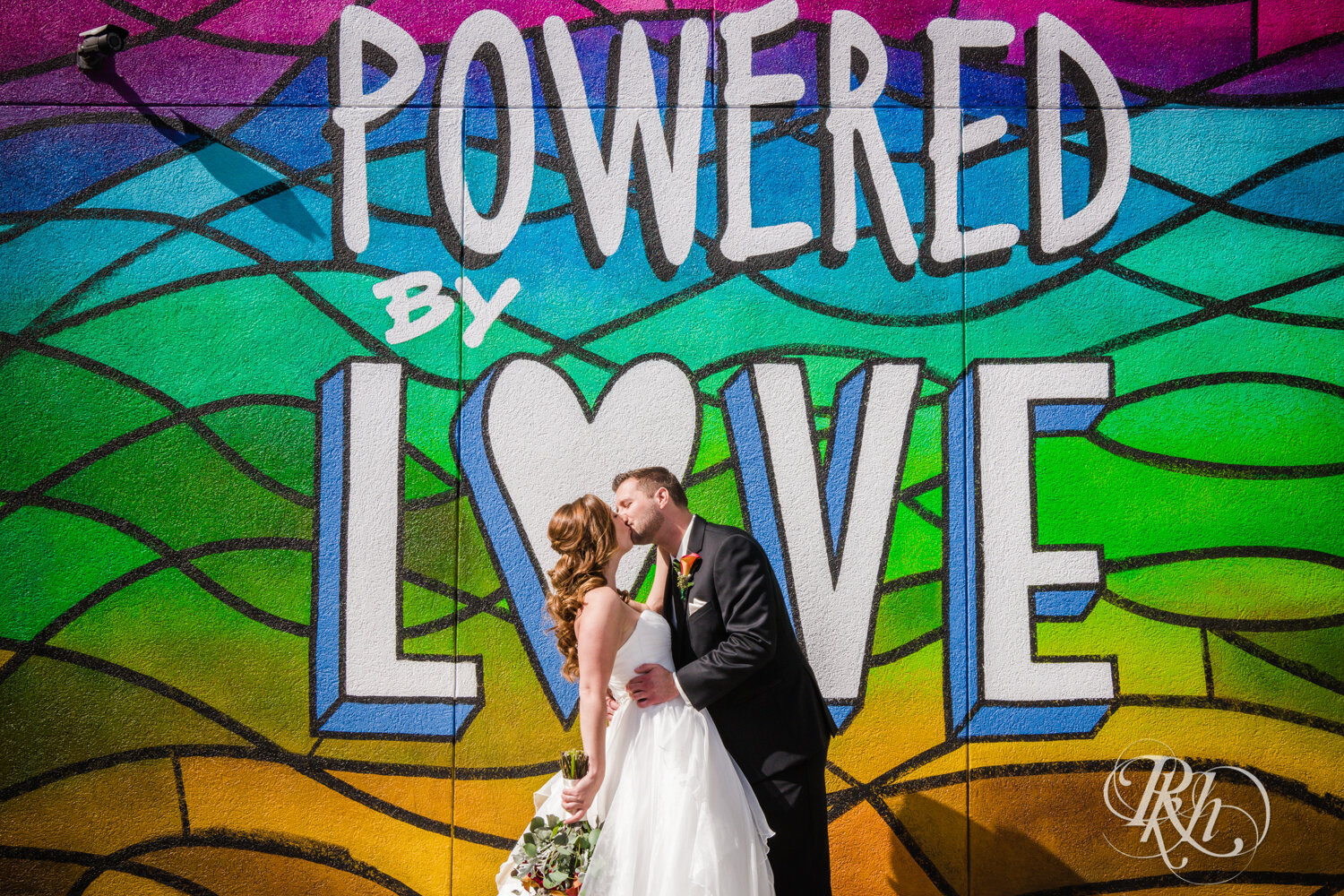 Bride and groom kissing in front of Powered by Love mural in Otsego, Minnesota on sunny wedding day.