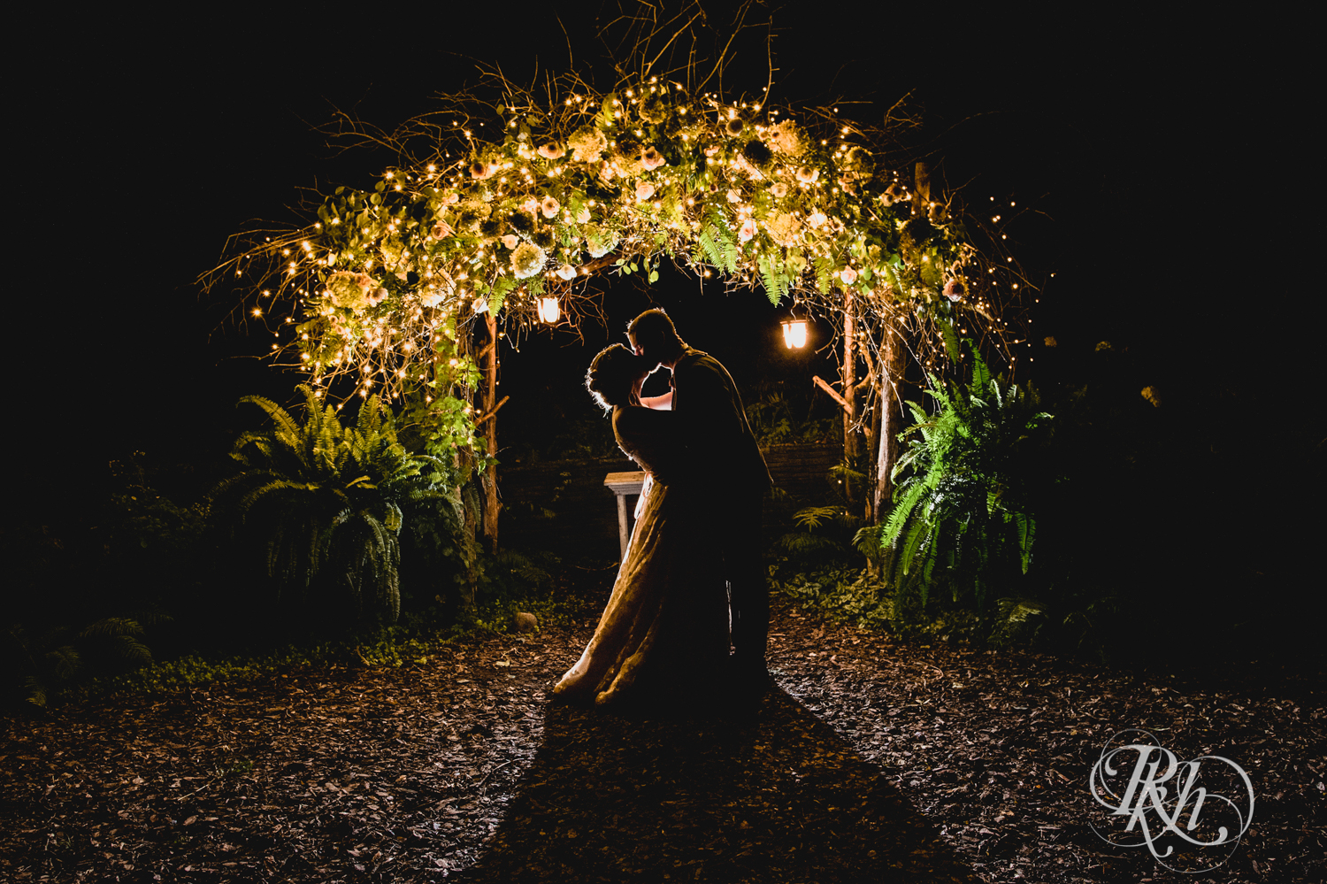 Bride and groom kiss at night in front of lights and flowers at Camrose Hill in Stillwater, Minnesota.