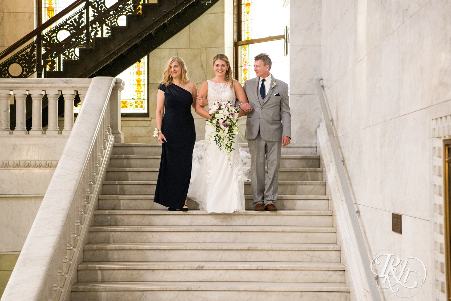 Bride walks down the stairs and aisle with parents at Minneapolis City Hall in Minneapolis, Minnesota.