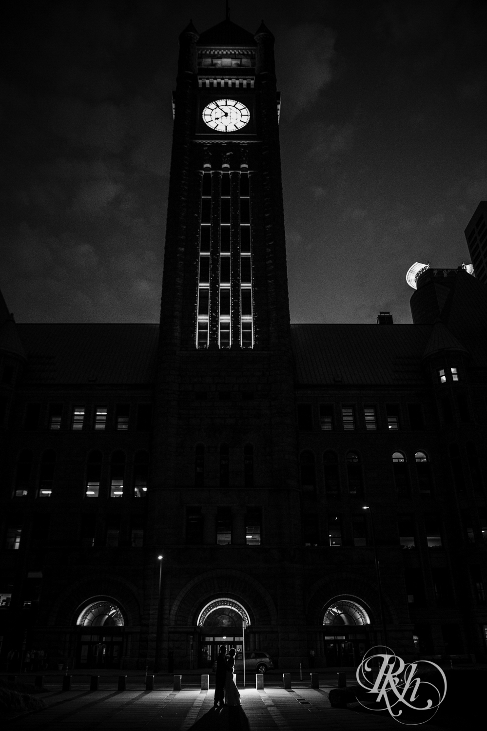 Bride and groom kiss at night under clock tower in Minneapolis City Hall in Minneapolis, Minnesota.