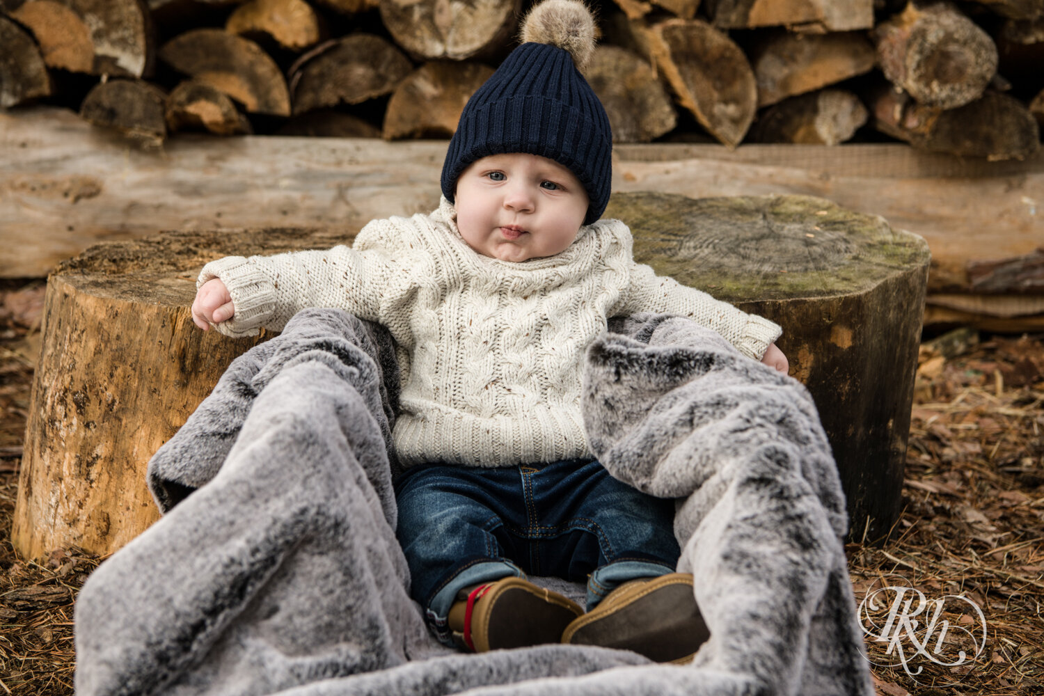 Baby in sweater and boots sits in sled in front of woodpile at Hansen Tree Farm in Anoka, Minnesota.