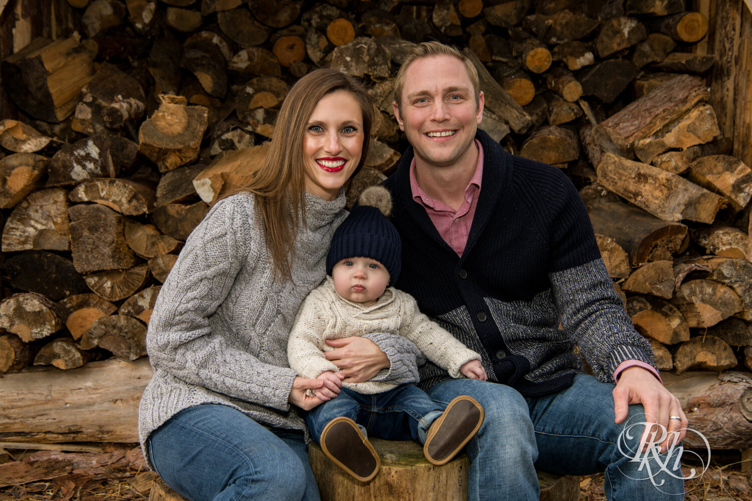 Man and woman hold baby and smile in front of woodpile at Hansen Tree Farm in Anoka, Minnesota.