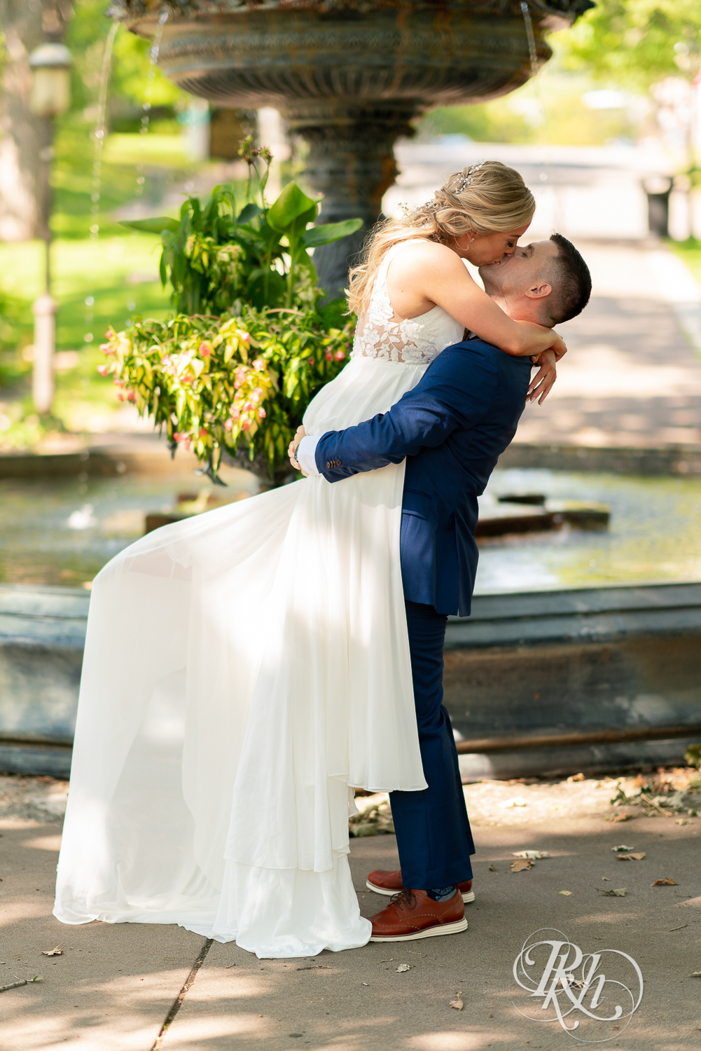 Groom lifts and kisses bride in front of fountain at Irvine Park in Saint Paul, Minnesota.