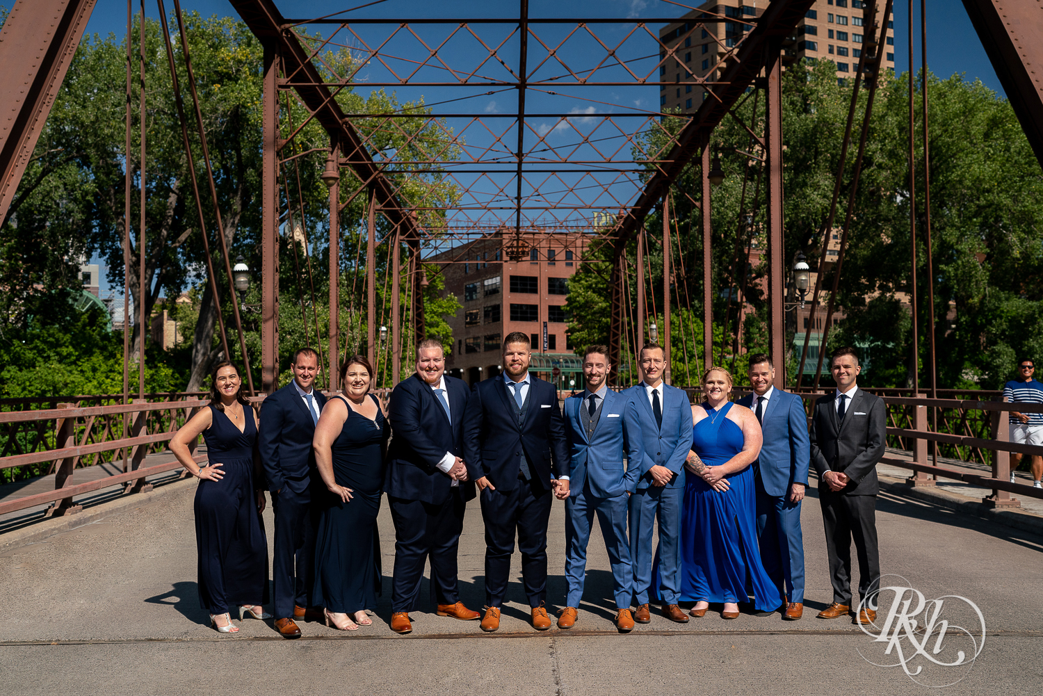 Gay wedding party dressed in blue suits and dresses smiling on bridge in Nicollet Island in Minneapolis, Minnesota on a sunny day.