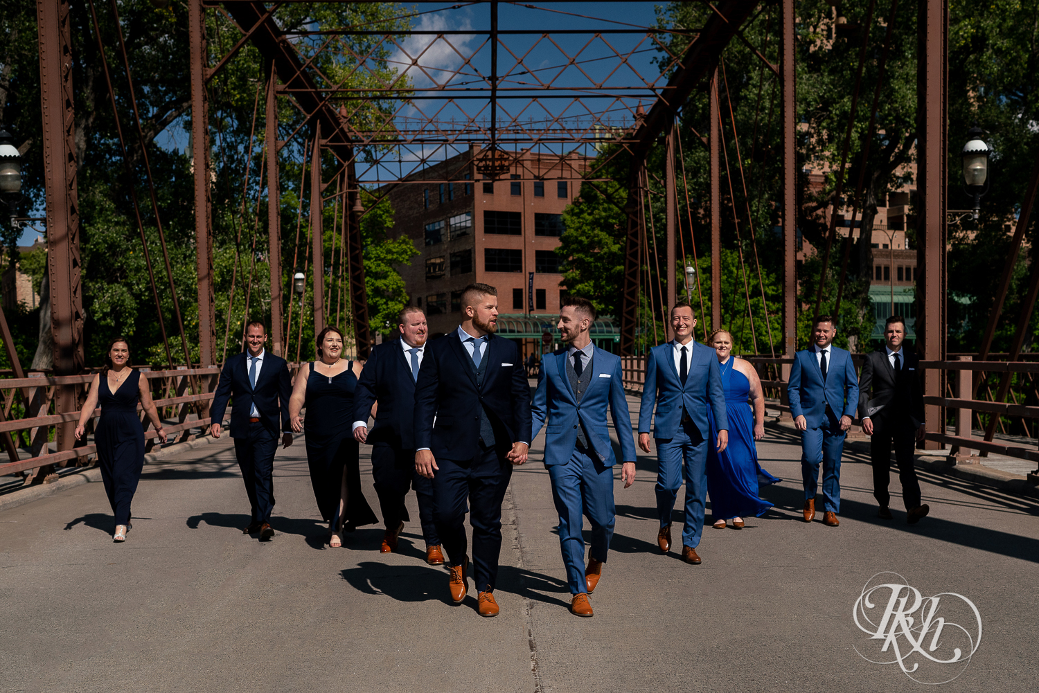 Gay wedding party dressed in blue suits and dresses walking on bridge in Nicollet Island in Minneapolis, Minnesota on a sunny day.