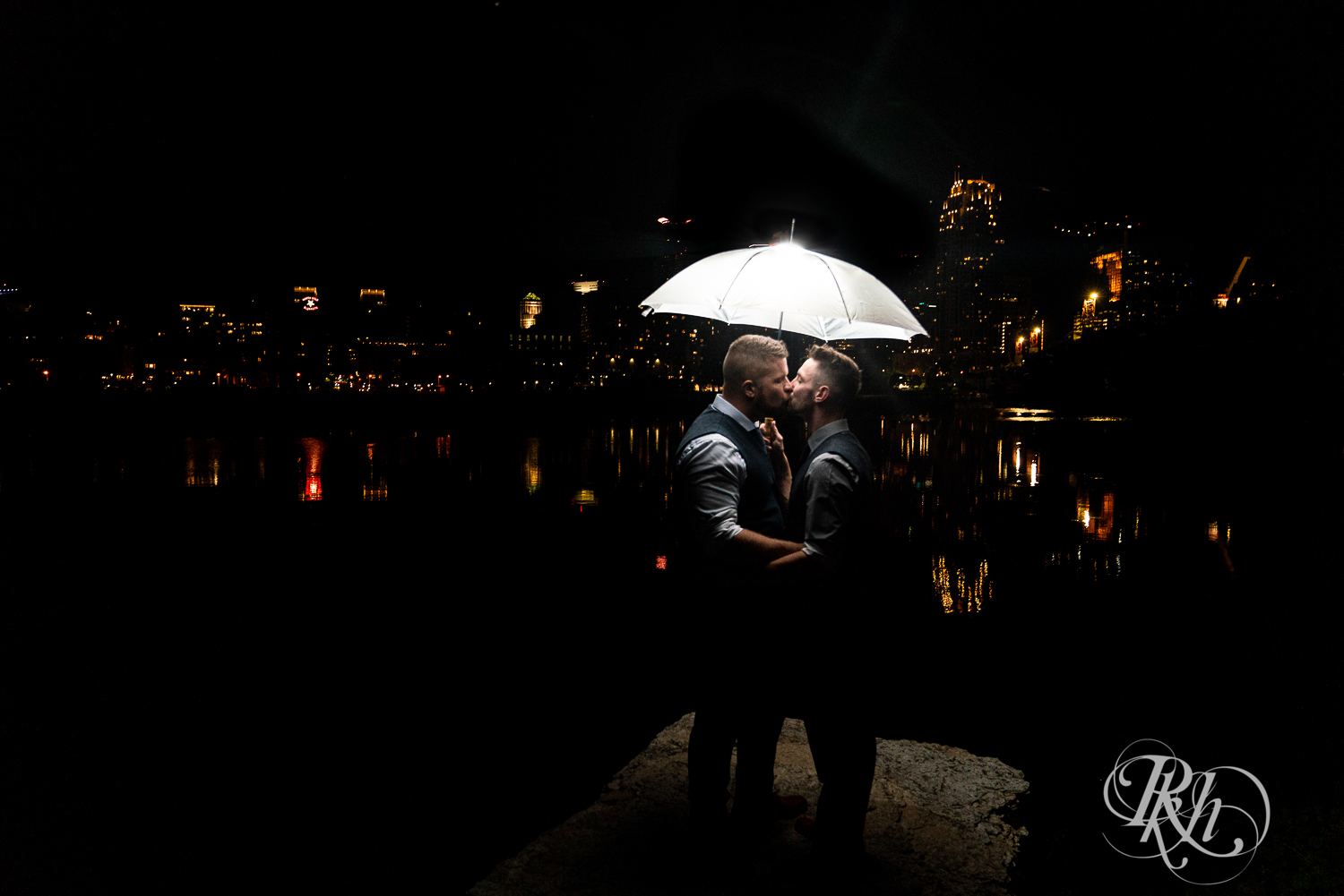 Gay grooms kiss under umbrella at night in Minneapolis, Minnesota with skyline in the background.