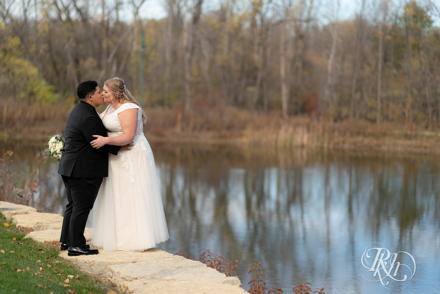 Lesbian brides kiss in fall leaves by pond at the Eagan Community Center in Eagan, Minnesota.