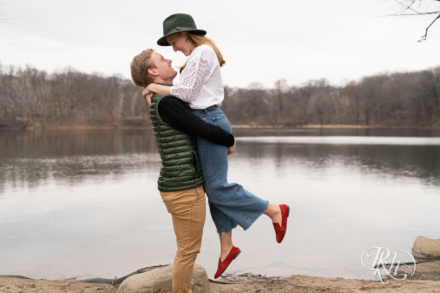 Man lifts woman dressed in jeans and a green hat in front of the lake in Eagan, Minnesota.