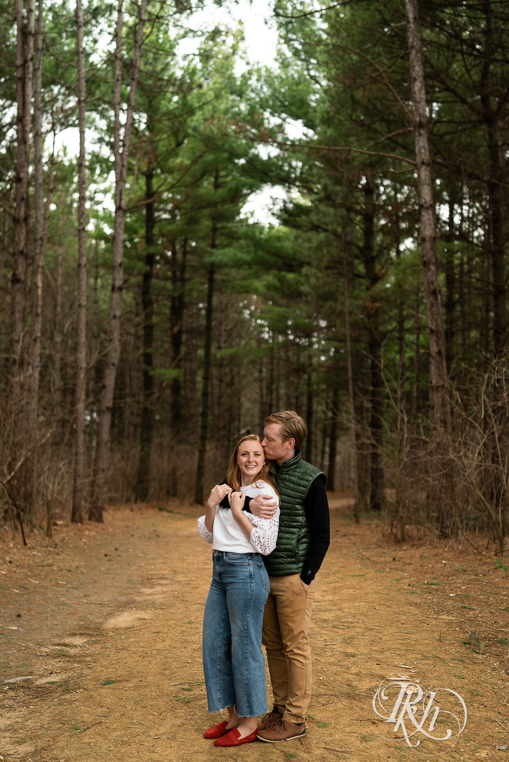 Man and woman dressed in jeans and a kiss in the woods in Eagan, Minnesota with pine trees in the background.