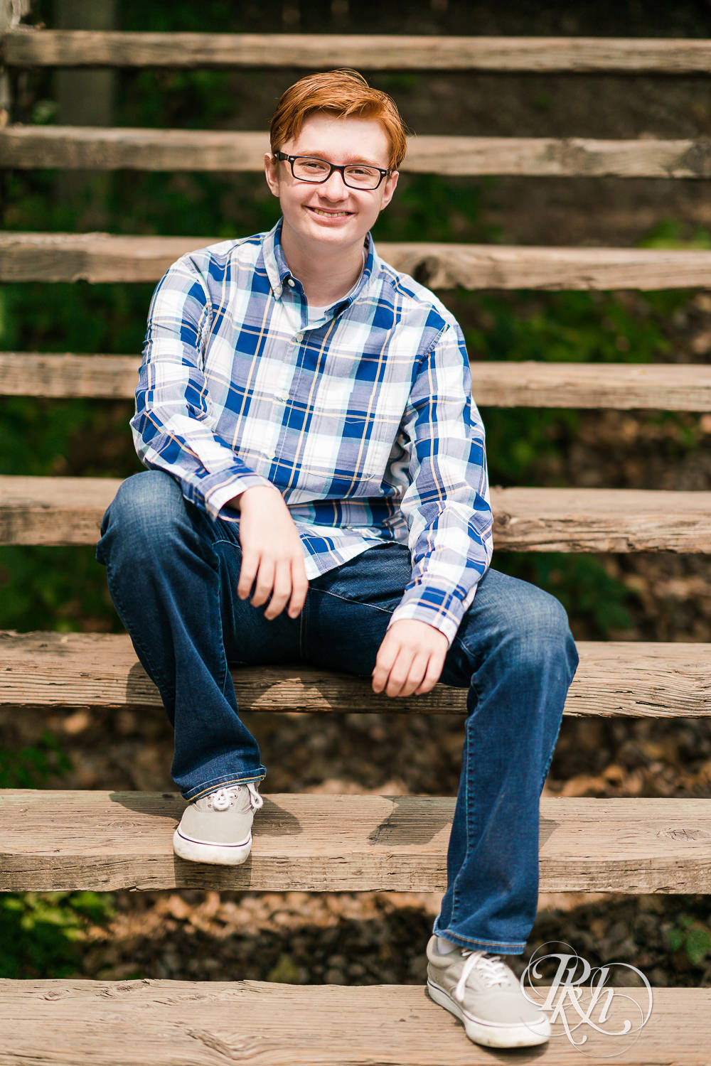 Redheaded boy in glasses and flannel poses on stairs for senior pictures in Minneapolis.