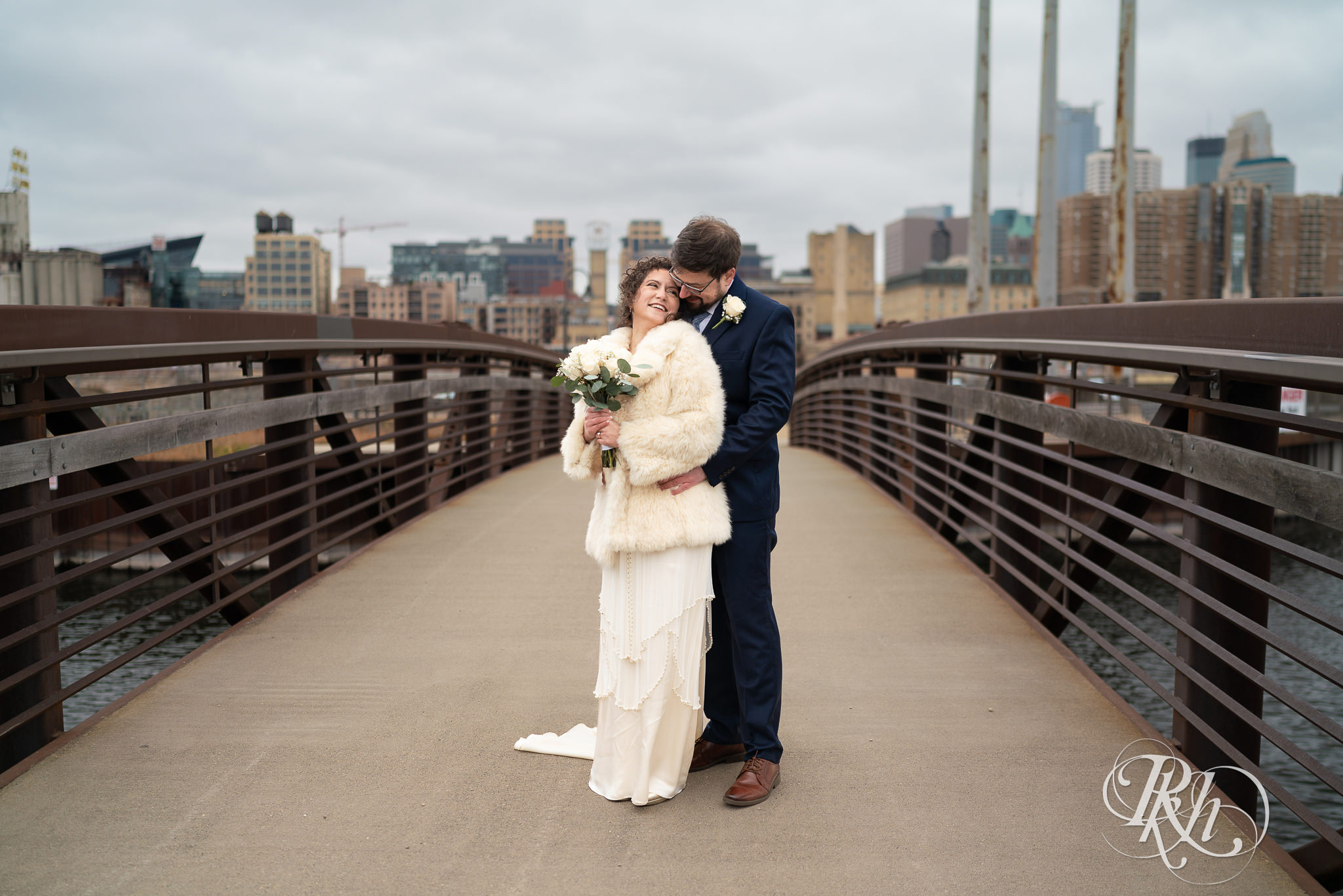 Bride and groom hugging and looking at each other on bridge with Minneapolis skyline in the background.