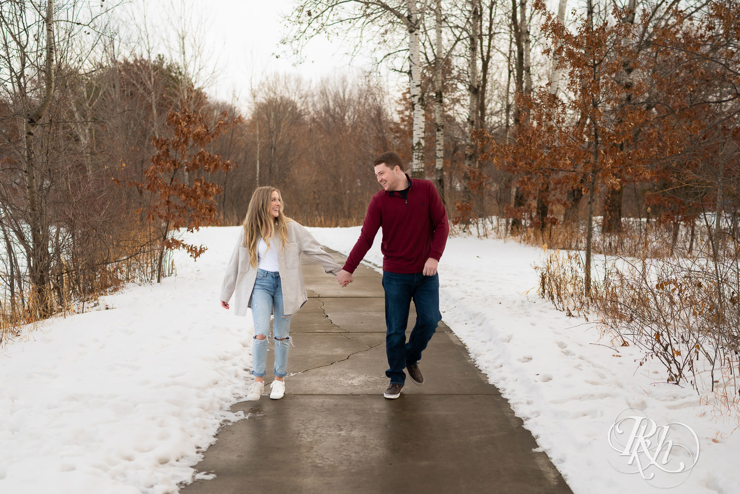 A couple walks down the snowy path in a park smiling at one another.