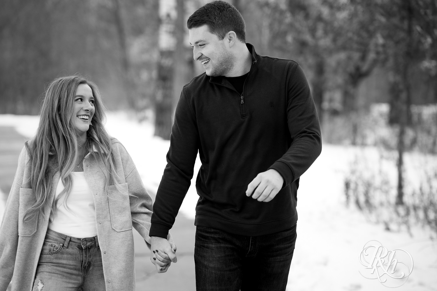 A couple walks down the snowy path in a park smiling at one another. The photo is in black and white.