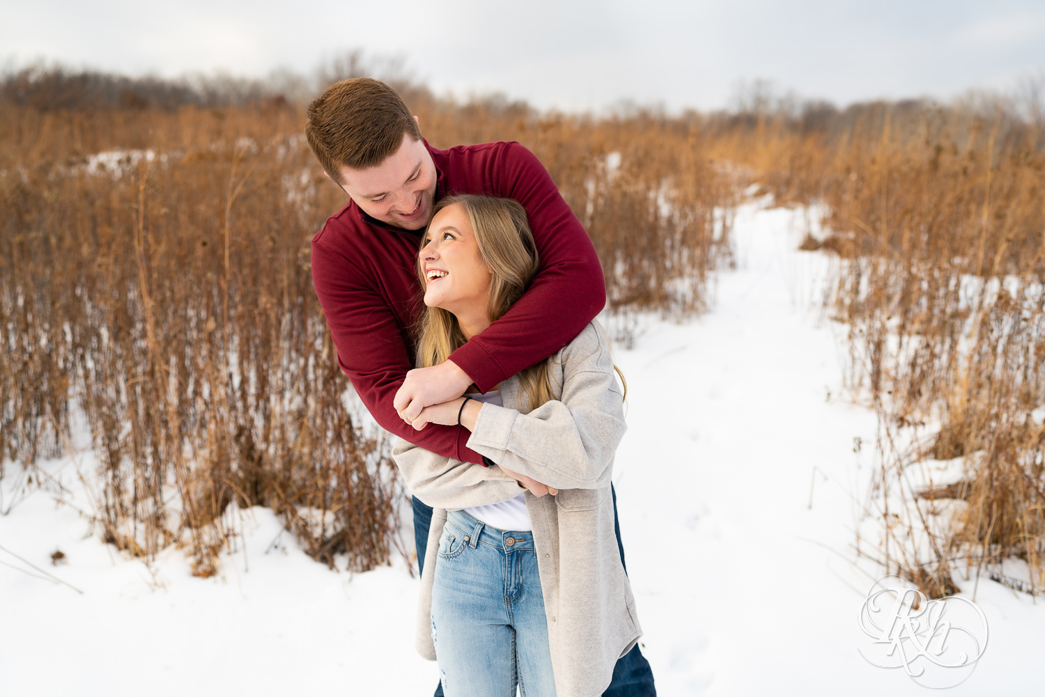A couple stands in the snow hugging and smiling at one another.
