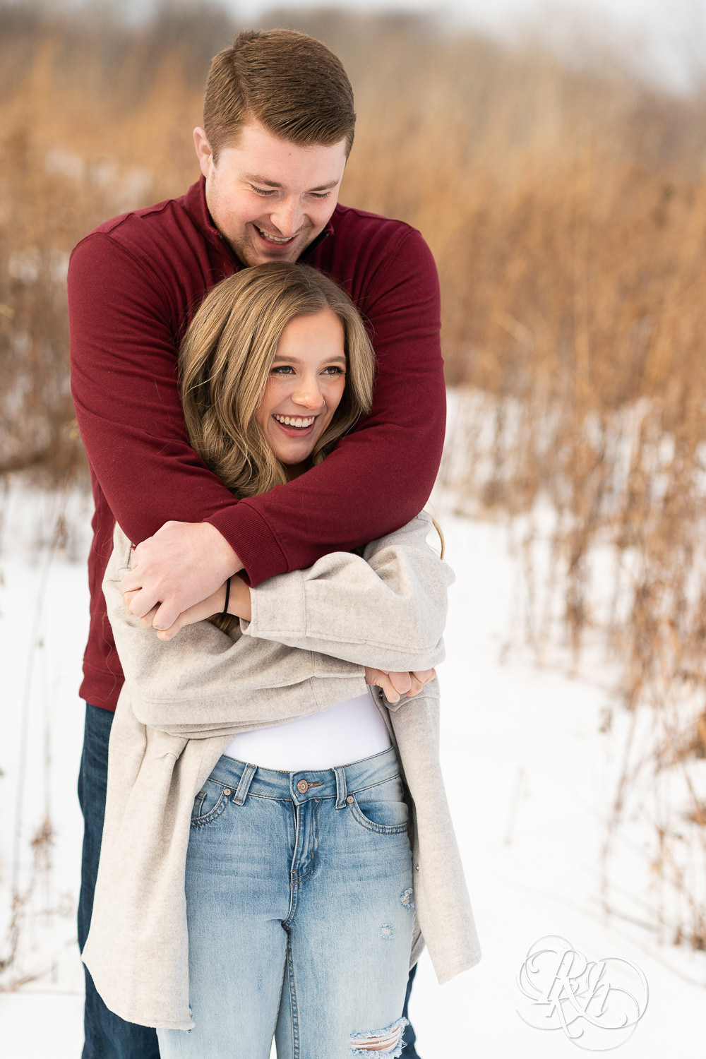 A couple stands in the snow hugging and smiling at one another.