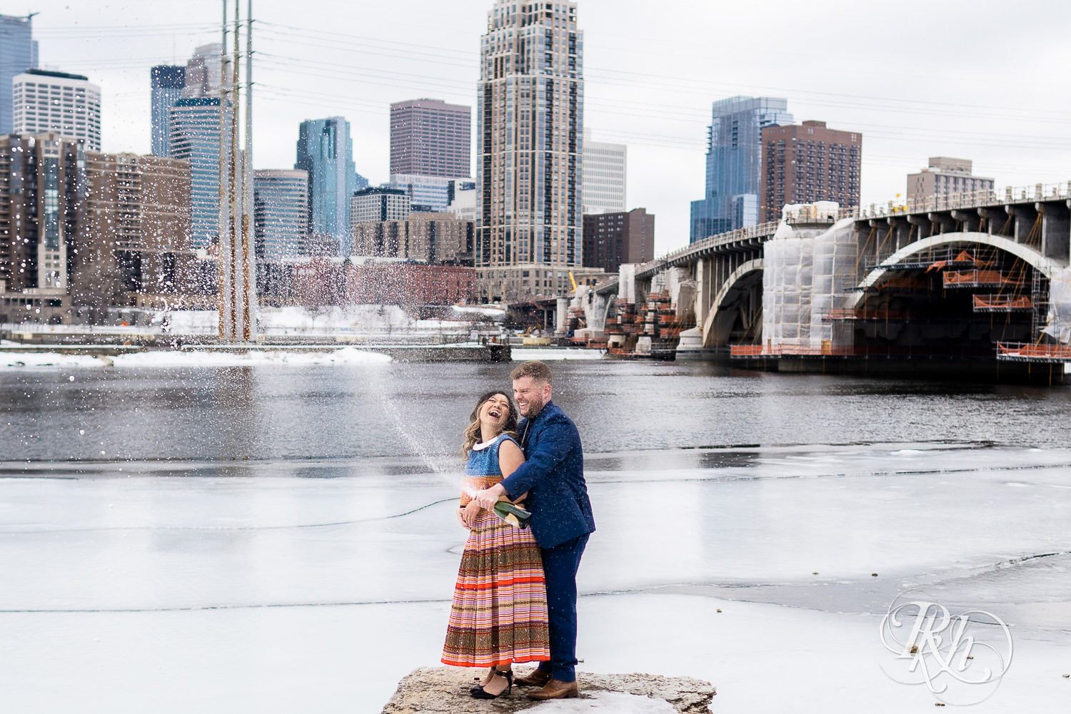 Man and Hmong woman spray champagne in Saint Anthony Main in Minneapolis, Minnesota with city in the background.