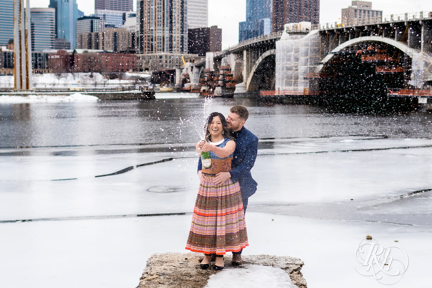 Man and Hmong woman spray champagne in Saint Anthony Main in Minneapolis, Minnesota with city in the background.