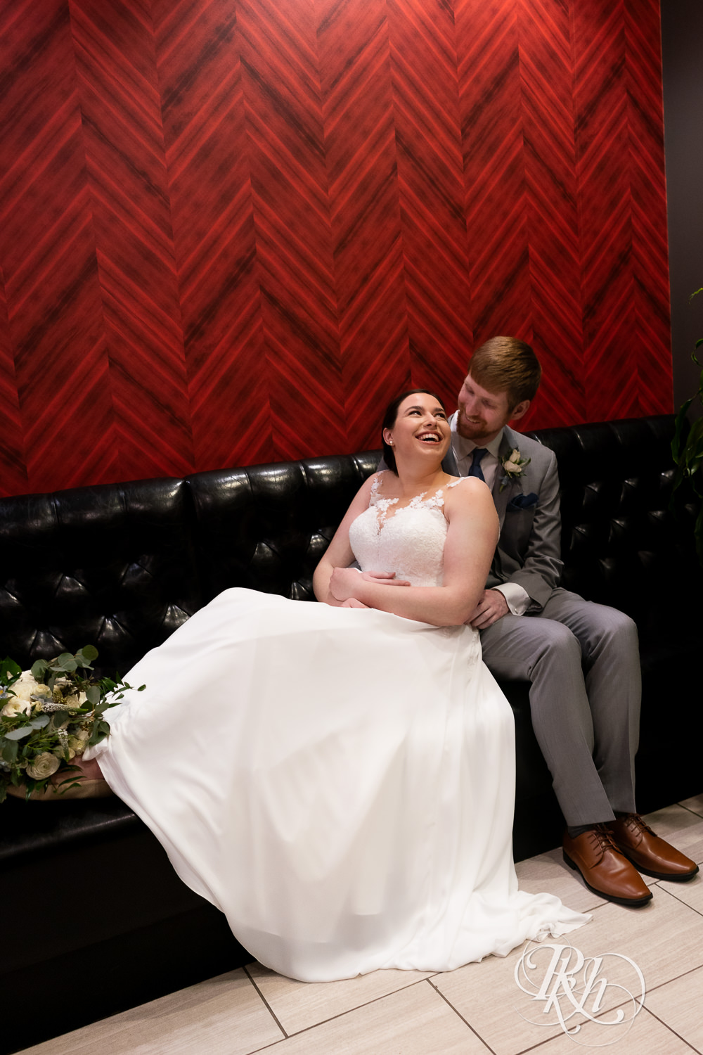 Bride and groom smile on couch by red wall at Doubletree Hilton Saint Paul in Saint Paul, Minnesota.